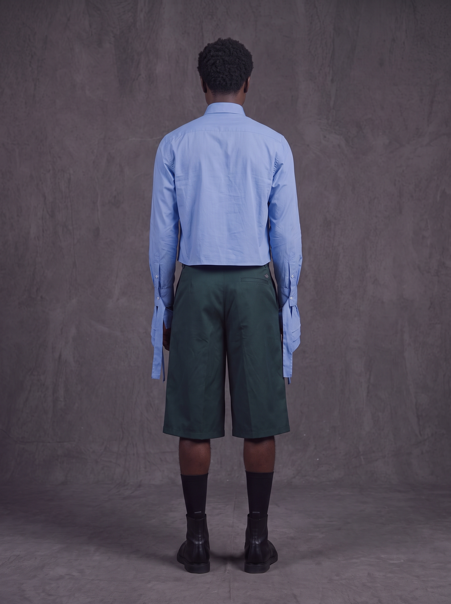 A model viewed from the back wearing a light blue long-sleeve button-up shirt, dark green knee-length shorts, black socks, and black boots, standing against a textured gray studio backdrop.