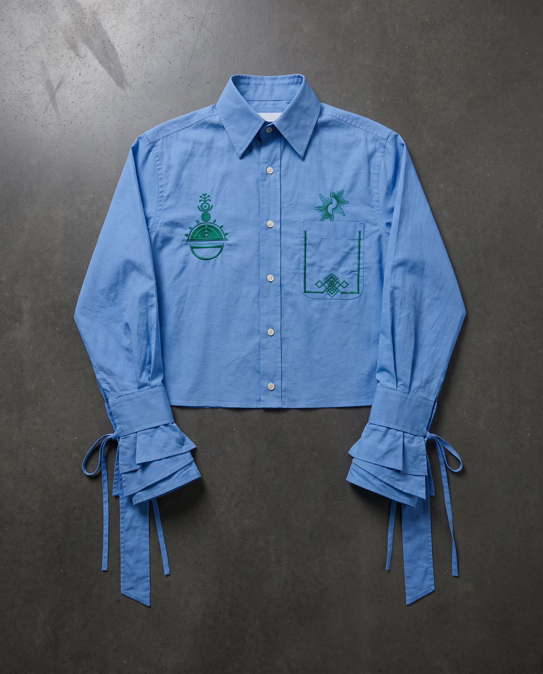 A light blue cropped button-up shirt with green embroidered details, featuring ruffled tie-back cuffs, laid flat on a dark textured background.
