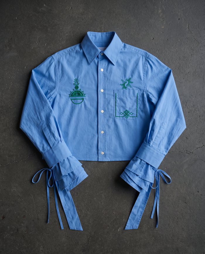 A light blue long-sleeve button-up shirt laid flat on a dark gray textured surface. The shirt features green embroidered graphics on the chest, including a palm tree and a geometric emblem, with matching blue fabric ties at the cuffs.