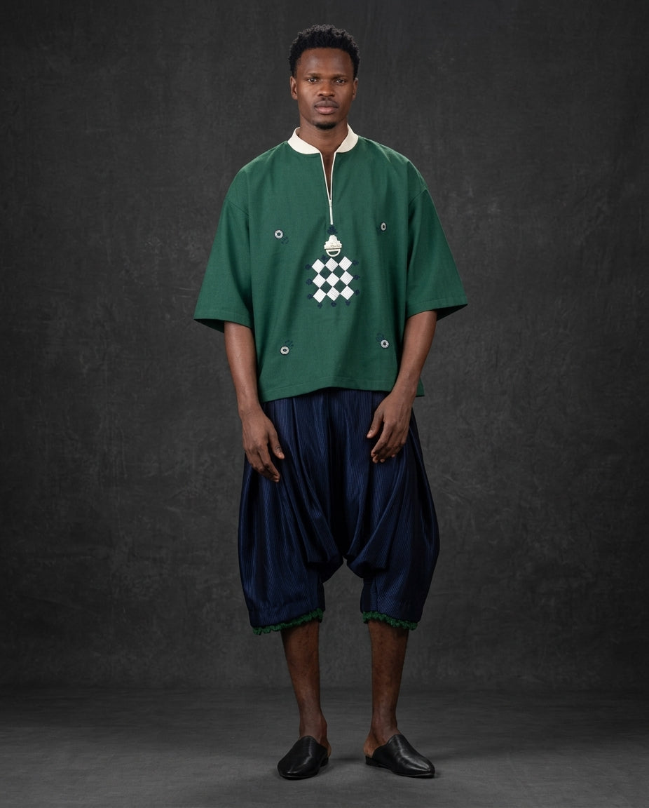 A man modeling a green short-sleeve tunic with a white geometric chest pattern and dark navy harem-style pants against a dark gray studio backdrop.