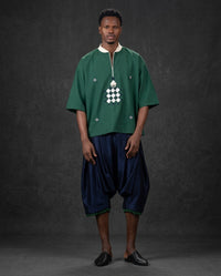 A man modeling a green short-sleeve tunic with a white geometric chest pattern and dark navy harem-style pants against a dark gray studio backdrop.