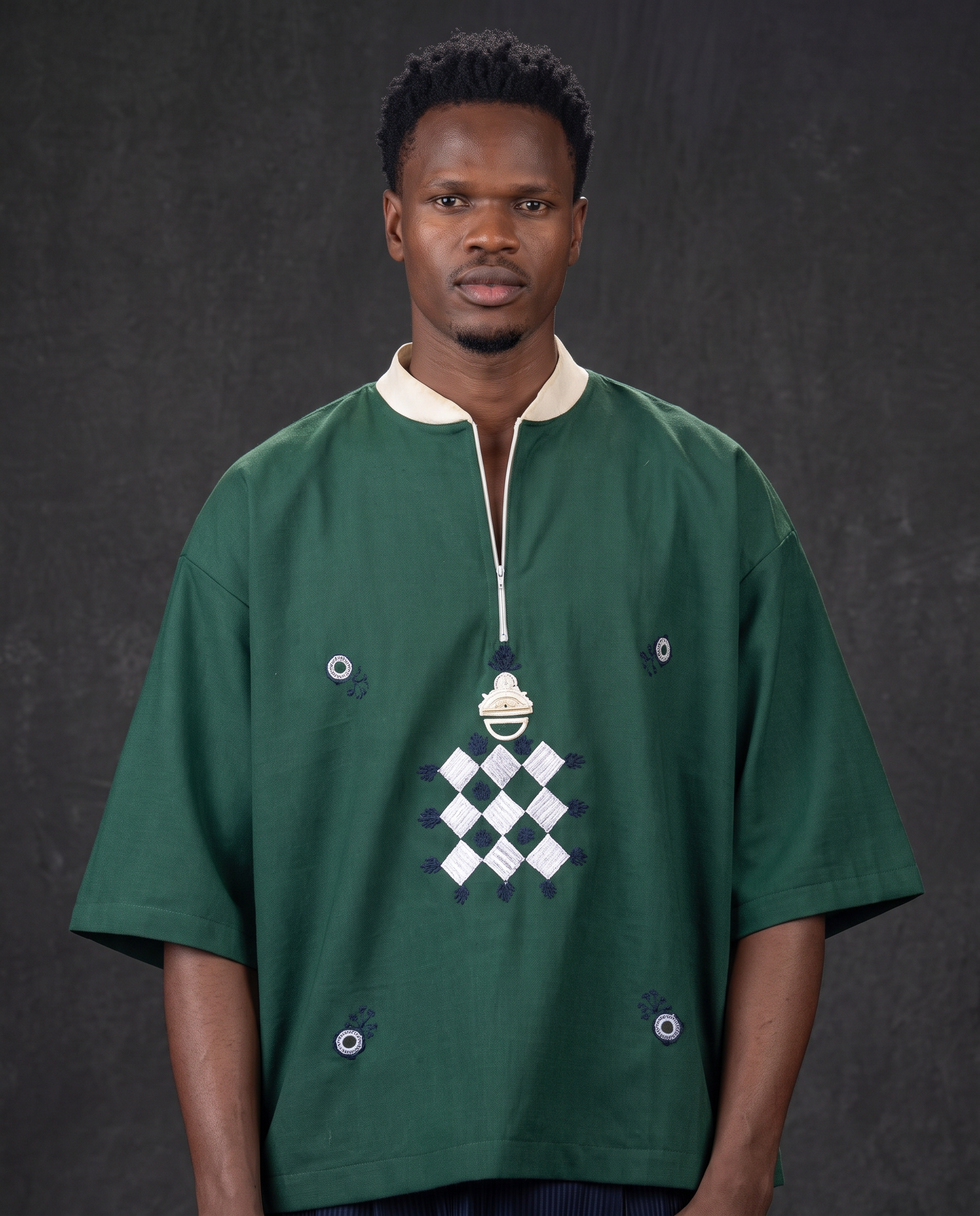 A man wearing a dark green traditional African shirt with a white collar, white zipper placket, and a central white and purple geometric diamond pattern, set against a dark gray textured background.
