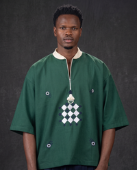 A man wearing a dark green traditional African shirt with a white collar, white zipper placket, and a central white and purple geometric diamond pattern, set against a dark gray textured background.