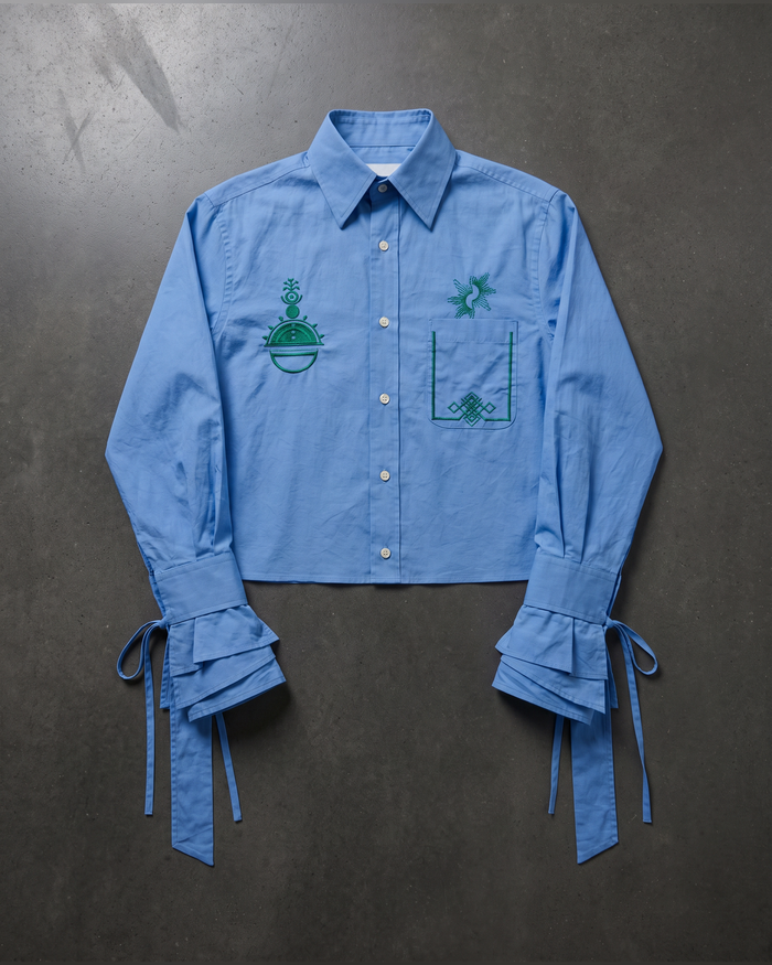 A light blue cropped button-up shirt with green embroidered details, featuring ruffled tie-back cuffs, laid flat on a dark textured background.