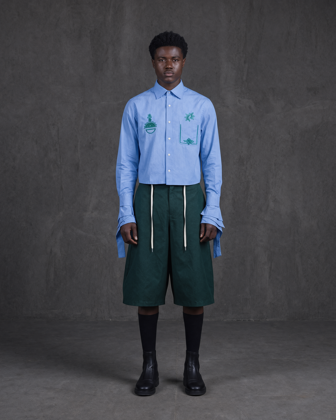 A model stands against a dark gray studio backdrop wearing a light blue long-sleeve button-up shirt with green graphic prints on the chest, dark green shorts with white drawstrings, black socks, and black boots.