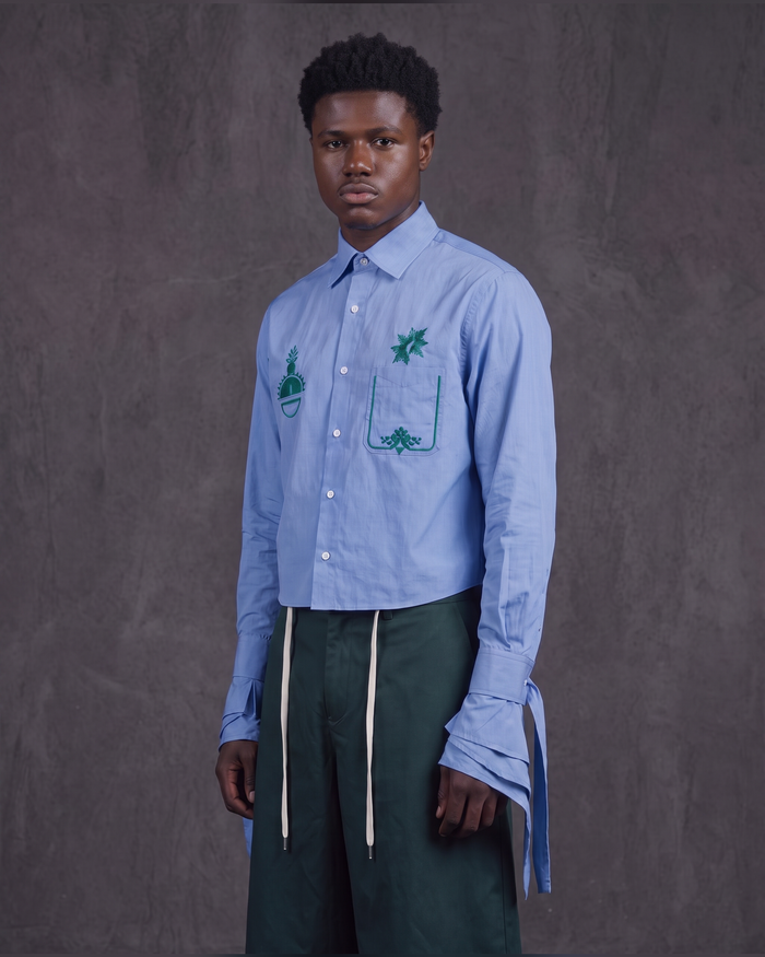 A male model stands against a textured gray studio backdrop wearing a light blue long-sleeve button-up shirt with green embroidered pocket details and dark green drawstring trousers.