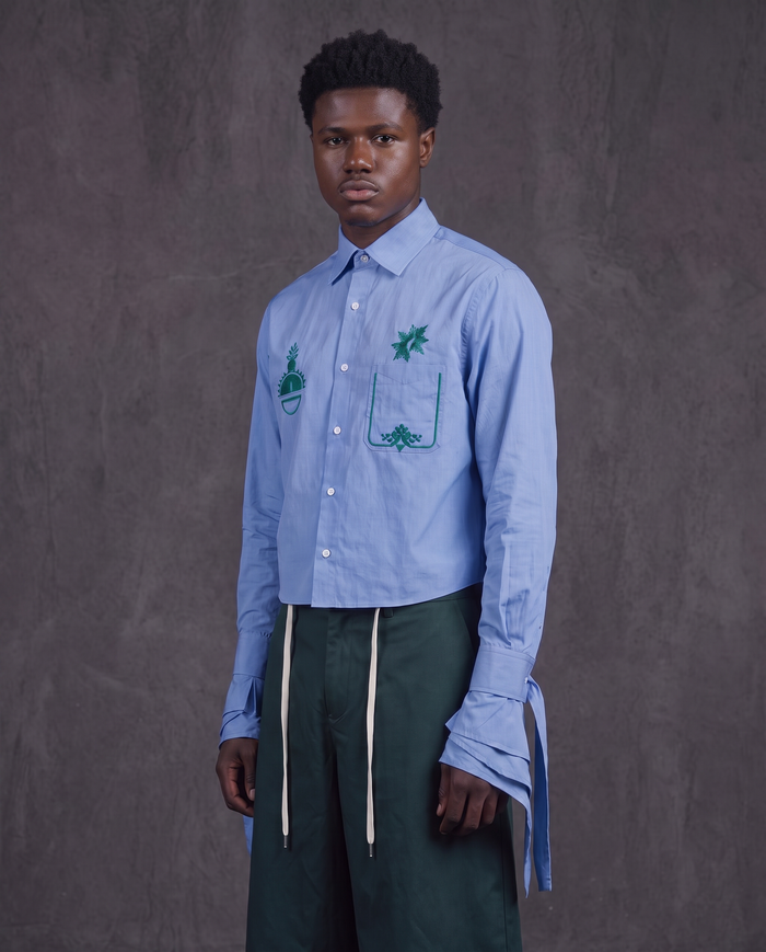 A male model stands against a textured gray studio backdrop wearing a light blue long-sleeve button-up shirt with green embroidered pocket details and dark green drawstring trousers.